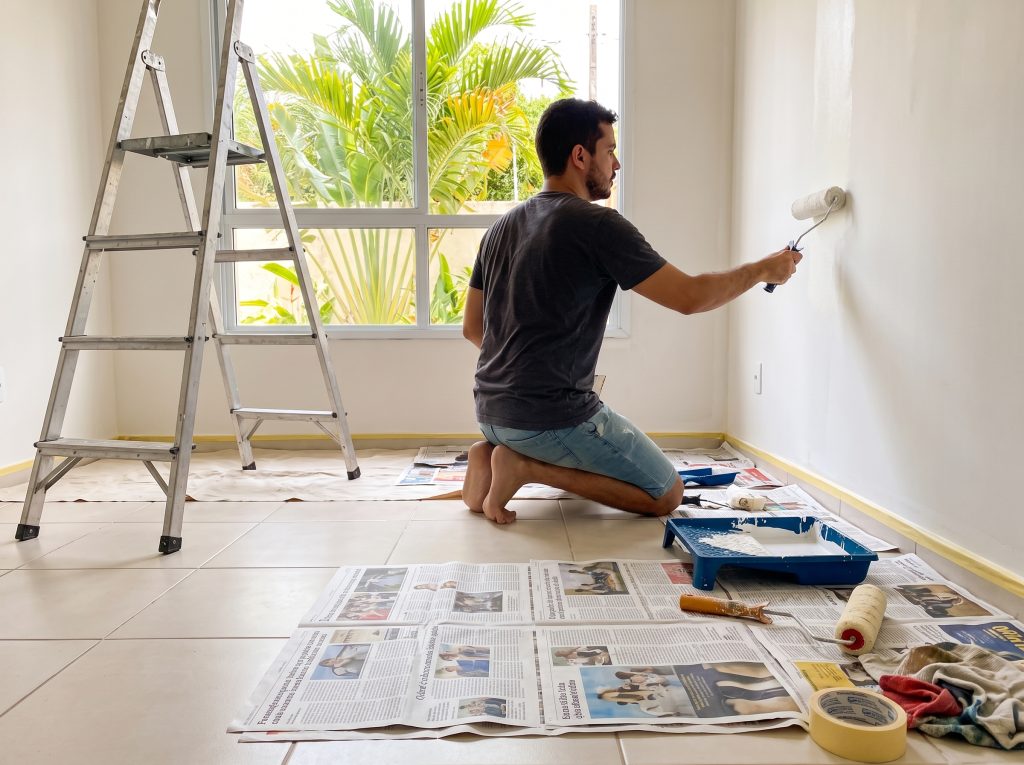 Homem jovem pintando parede interna com rolo e tinta para parede clara em casa, com materiais de pintura como bandeja e fita sobre jornais no chão, escada de alumínio ao lado e janela com vista para plantas tropicais.